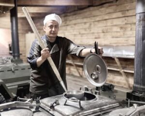 – Потом привык, наладил работу, – продолжает «Рафит». – Готовлю на батальон завтрак, обед, ужин. Есть повара, которые помогают. В мирной жизни военнослужащий к плите не подходил, но армейская служба напомнила о навыках, приобретённых в юности. – Учли, что службу по призыву проходил поваром, вот и назначили, – скромно объясняет он. Сейчас его должность – создатель фронтовых кулинарных шедевров, как с тёплой иронией говорят сослуживцы. В его распоряжении четыре машины полевой кухни. – Обучение на них проходили с первых дней, – делится повар. Подъём в три ночи – и процесс начинается. – Запускаю печи, истопник с дровами помогает, я поджарку готовлю, мясо разделываю, – описывает он свой обычный день. Уже к пяти утра, когда военнослужащие собираются на полигон, их ждёт горячий завтрак. К полудню готов ужин, обед – к пяти вечера, точно к возвращению личного состава с полигона. Работа на кухне кипит безостановочно. Только к 20.00 наступает относительное затишье. – В конце рабочего дня наводим порядок: моем посуду, инвентарь возвращаем на места, проверяем электричество, оборудование, – рассказывает старший повар. – Подготавливаем кухню к следующему дню, набираем воду. Только после этого уходим отдыхать. – Раскладка приходит на неделю, по ней мы и работаем, – объясняет он. – Сколько и каких продуктов заложить – всё расписано. Ежедневно кухня получает суточную норму продуктов от коман­дира взвода продовольственного обеспечения, который занимается их получением со склада и распределением. За качество поставок «Рафит» не волнуется. – Проблем с продуктами нет, – говорит он с гордостью. – Если вложить душу, можно приготовить что угодно. Главное, чтобы все были сыты и довольны. Рацион солдата таков, чтобы обеспечить сытость и запас энергии. На завтрак отварное мясо, каша, чай, масло, хлеб, печенье, шпик и салаты. А обед – основа основ. – Первое блюдо – суп. Всегда разный: гороховый, рассольник, борщ, щи. Второе – обязательно каша с мясом. Третье – соки натуральные, – рассказывает старший повар. «Рафит» особо заботится о питьевом режиме стоящих на довольствии. – Стараюсь выдавать как можно чаще чай и соки, потому что сейчас пора жары. Парни на полигоне, им требуется больше жидкости, – подчёркивает он. Вместе с супом и кашей идут салаты, хлеб, печенье, вафли. Ужин – гарнир с мясом. Особое место в меню старший повар уделяет горячему. – Крайне важная часть рациона – это суп, – говорит он, помешивая ароматное варево в огромном котле. – Понимаете, одним сухим пайком всегда сыт не будешь. Конечно, он незаменим на переднем крае, когда просто нет возможности обеспечивать военнослужащих свежим и горячим питанием. На родине «Рафита» ждут жена и трое детей. Ирония судьбы: дома он к плите не подходит. – Когда приезжаю в отпуск, вообще не готовлю. Хватает на службе, – с улыбкой говорит он и добавляет, что в еде непривередлив. – Супругу это очень радует. Потому что чего-то конкретного никогда не прошу. Без громких слов и претензий на гурманство старший повар «Рафит» каждый день совершает свой подвиг. Его полевая кухня – не просто место приготовления пищи. Это островок тепла и заботы среди военных будней. Где вкусный суп, сваренный уже чуть ли не с закрытыми глазами, но с открытым сердцем, становится не просто едой, а воспоминанием о доме, напоминанием о мирной жизни и является той самой силой, что помогает солдатам идти вперёд. Его ложка такое же оружие победы. Группировка войск «Центр»